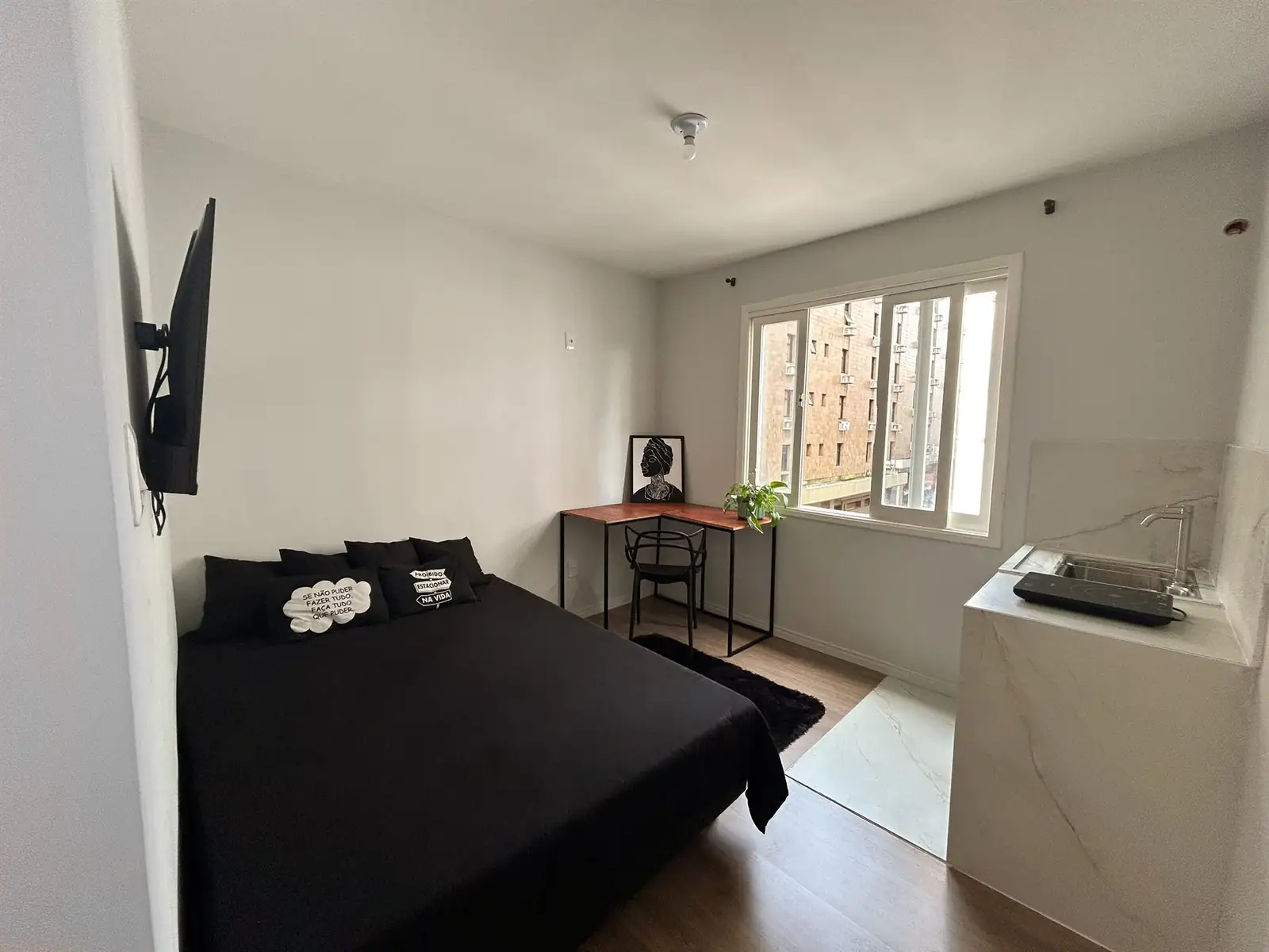 Estúdio com cama de casal, TV na parede, mesa junto à janela e cozinha no canto