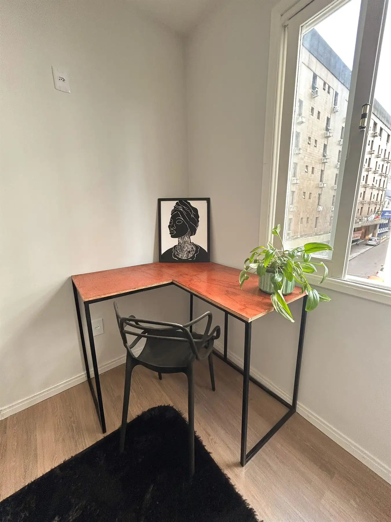 Mesa de trabalho em L no canto do estúdio, com cadeira e planta junto à janela